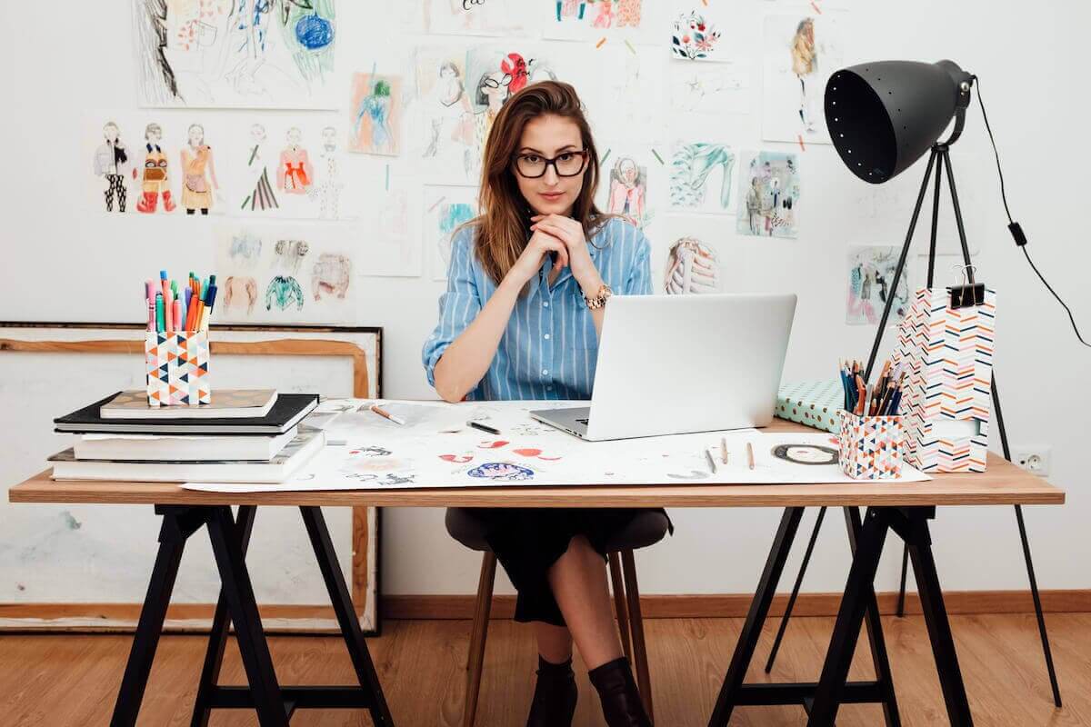 A woman wearing glasses sits at a desk with a laptop and papers.