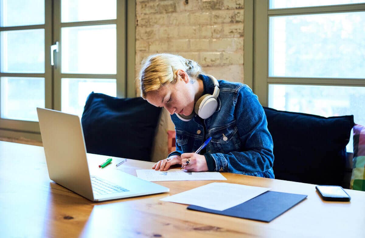 Female student wearing headphones writes with pen at table with laptop and mobile phone nearby