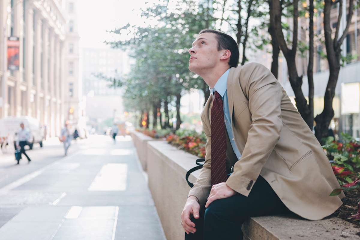 Young man in sport coat and tie sits on low wall in financial district