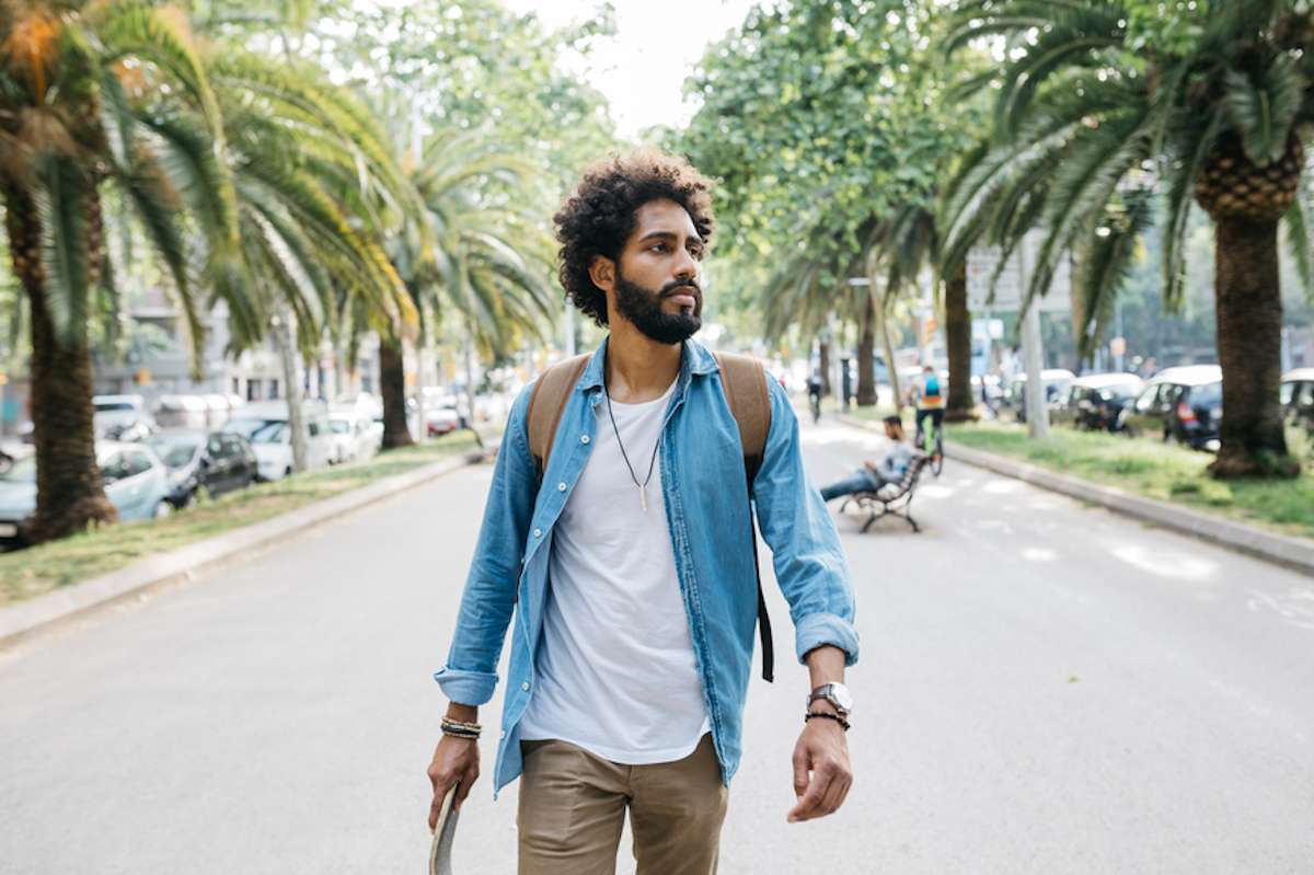 Bearded man in denim shirt walks outside carrying a skateboard and wearing a backpack