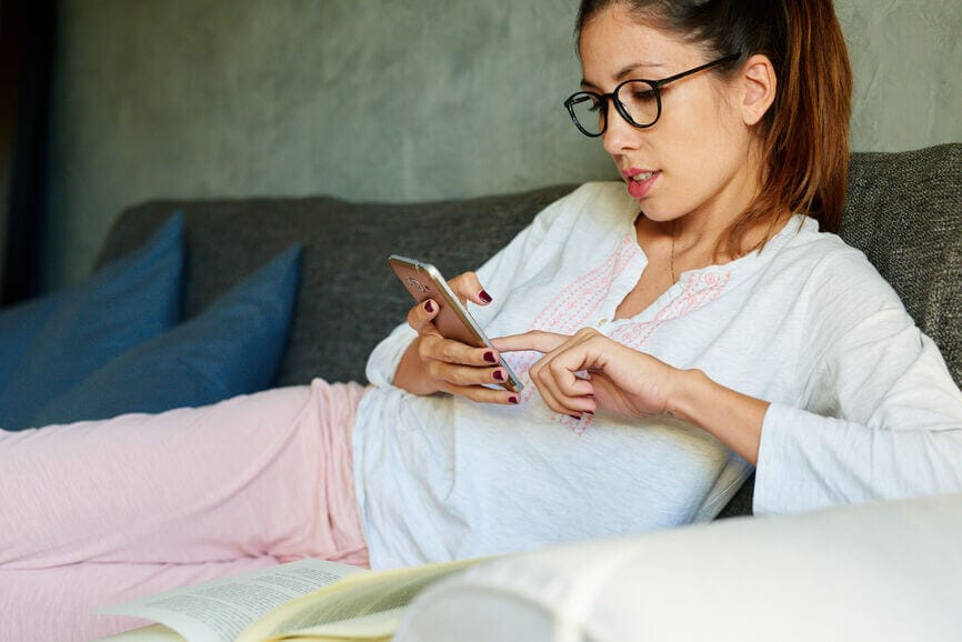 Young woman wearing glasses scrolling through her phone on her couch.