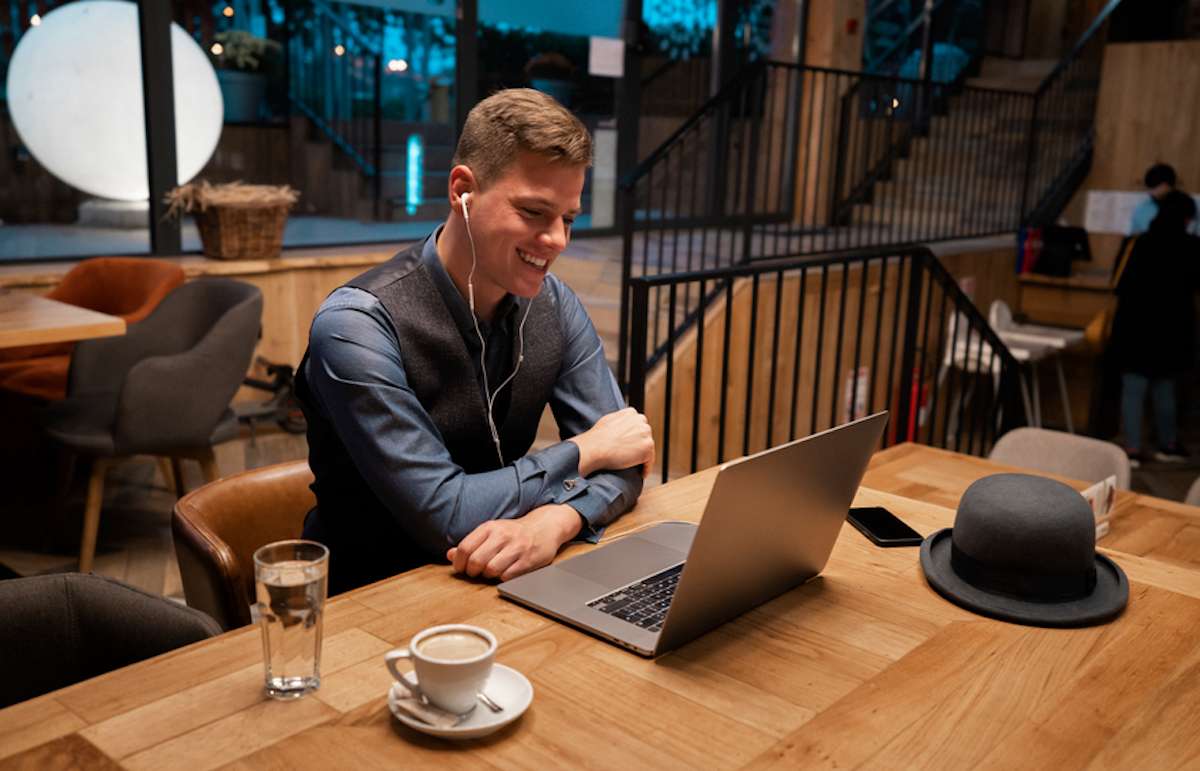 Un joven sentado en un café sonríe mientras trabaja en su computadora portátil.