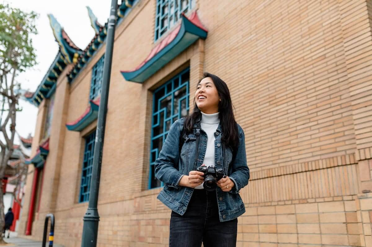 A smiling Asian woman walks down a street with her camera around her neck.