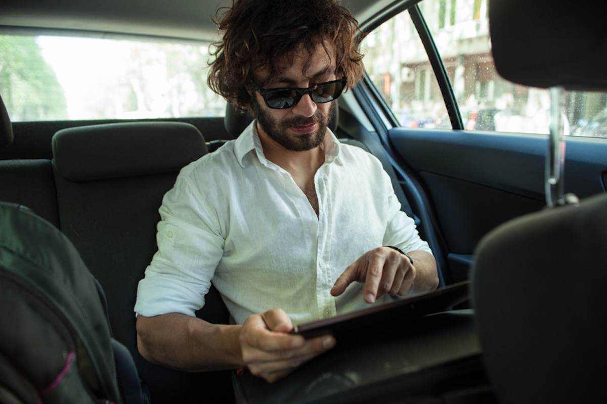 Casually dressed young man works on his tablet in the back seat of a car