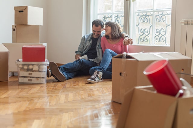 A couple sitting together on the floor of their living room, surrounded by moving boxes.