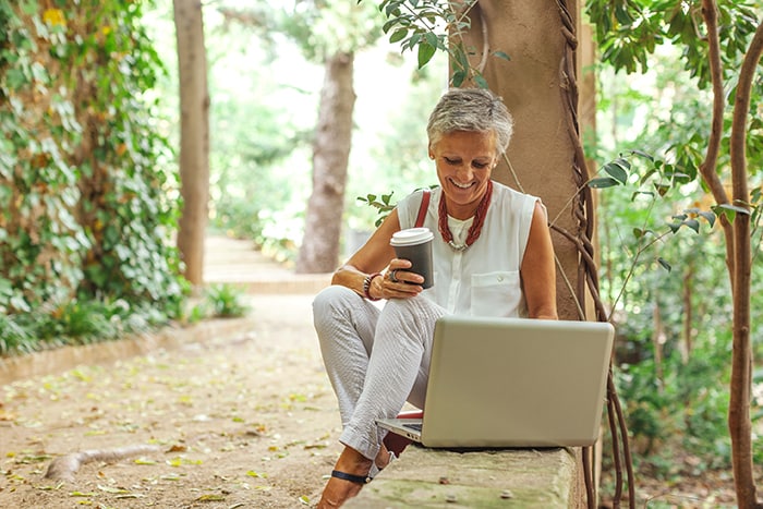 Person in forest looking at laptop while smiling with coffee
