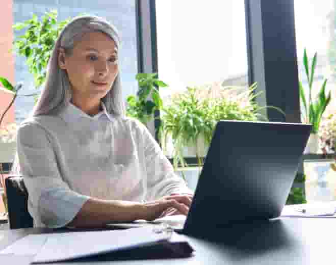 Asian woman in white blouse working on dark grey laptop in bright sunlit office