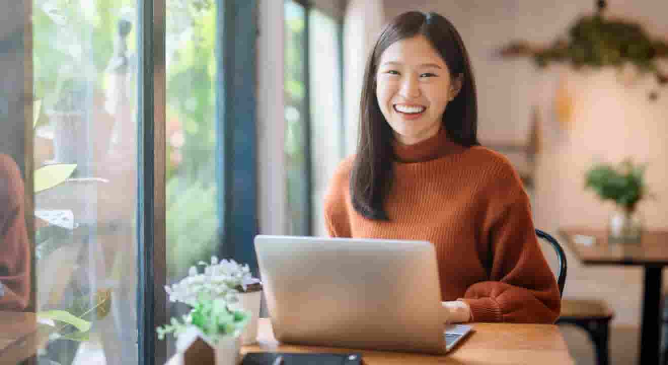A woman in an orange sweater and looking up from her laptop with a smile.