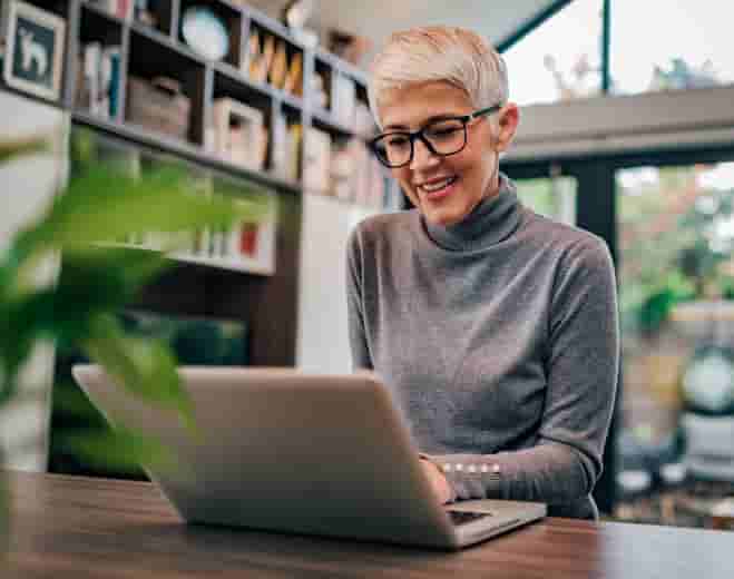 Older woman in grey sweater smiles while working on sleek laptop