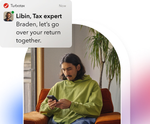 A man in his mid-20s wearing a green hoodie sits on his couch looking at his phone. He sees a message from Libin, a TurboTax expert. The message says: Bradon, let’s go over your return together.