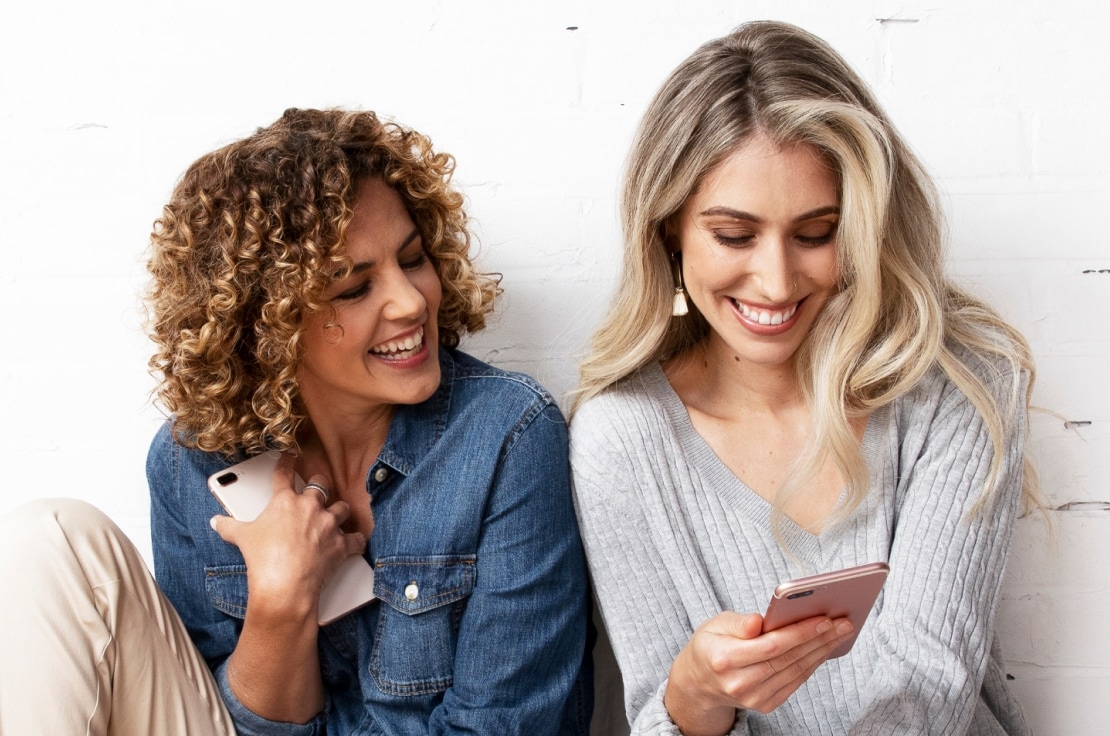 a blonde woman and a curly-haired brunette women smiling and looking at their cell phones